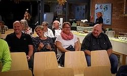 Eine Gruppe von Erwachsenen, die in Stühlen in einem Restaurant sitzen, möglicherweise bei einer Veranstaltung. In der Mitte befinden sich zwei Frauen mit Brille und Schals. Rechts steht ein Kühlschrank.