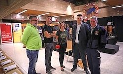 Fünf Personen stehen in einem Raum und lächeln. Drei von ihnen sind Männer und zwei sind Frauen. Sie tragen alle Freizeitkleidung. Eine Frau hält eine rote Handtasche. Es gibt Stühle und einen Tisch im Raum.
