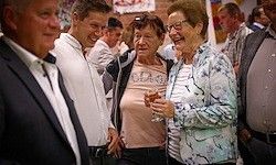 Eine Gruppe von Erwachsenen, sowohl Männer als auch Frauen, lächelt und steht zusammen. Eine Frau hält ein Glas Wein. Sie sind formell gekleidet, und der Hintergrund ist verschwommen mit mehr Menschen.