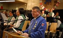 Ein Mann spricht an einem Podium und hält ein Buch in der Hand, während eine Band und ein Chor hinter ihm sitzen. Das Publikum und die Fahnen sind im Hintergrund.