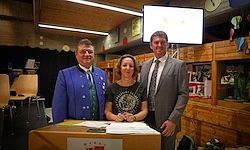 Drei Personen stehen hinter einem Tisch. Die Frau in der Mitte unterzeichnet ein Dokument. Zwei Männer stehen hinter ihr in Anzügen und Krawatten.