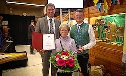 Drei Menschen stehen nebeneinander. Ein Mann hält einen Zertifikat und einen Blumenstrauß. Eine ältere Frau hält eine Medaille. Sie stehen vor einer Holzwand.
