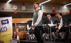 Ein Mann in einer grünen Weste steht lächelnd mit einem Mikrofon in der Hand auf der Bühne, mit einer Band, die verschiedene Instrumente spielt. Hinter ihm befindet sich ein gelbes Banner mit weißem Text.