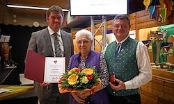 Drei Menschen stehen in einem Raum zusammen. Ein Mann hält ein Zertifikat, eine Frau eine Blumenbouquet und ein weiterer Mann eine Trophäe. Sie lächeln und scheinen sich bei einer Preisverleihung zu befinden.