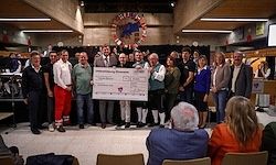 Eine Gruppe von Menschen steht um einen Mann, der einen großen Scheck in der Hand hält, umgeben von anderen Menschen in verschiedenen Kleidungsstücken, die in einem Saal mit Stühlen und einer Bühne stehen.