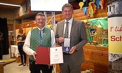 Zwei Männer stehen und halten ein Zertifikat und eine Medaille. Ein Mann trägt eine grüne Weste, der andere einen grauen Anzug. Sie sind in einem Raum mit Holzwänden und Dekorationen.