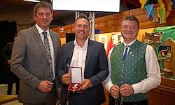 Drei Männer stehen in formeller Kleidung zusammen; einer hält eine Medaille und ein Dokument. Sie befinden sich in einem Raum mit Holzwänden und einem Hintergrund aus bunten Flaggen.