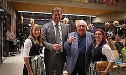 Vier Menschen posieren für ein Foto im Innenraum, zwei Frauen in traditioneller deutscher Kleidung, ein Mann in Anzug und ein Mann in Blazer, mit einer Menge im Hintergrund.