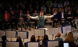 Ein Dirigent führt ein Orchester bei einer Aufführung an, mit einem großen Publikum, das zusieht und in Reihen auf Stühlen sitzt, während Musiker ihre Noten halten.