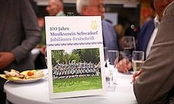 Eine weiße Karte mit dem Text '100 Jahre Musikverein Schwäbisch Jubiläums-Festschrift' steht auf einem Tisch mit Weinläsern und einem Teller Essen. Im Hintergrund sitzen verschwommen Menschen am Tisch.