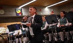 Ein Mann steht mit einem Mikrofon und hält ein Blatt Papier in der Hand. Hinter ihm sitzen eine Gruppe von Menschen in traditioneller Kleidung und spielen Instrumente. Der Raum ist gut beleuchtet, und es gibt Musikinstrumente und eine Uhr an der Wand.