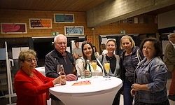 Eine Gruppe Erwachsener, einige mit Weinglas in der Hand, steht um einen weißen Tisch herum. Dahinter zeigt eine Wand verschiedene gerahmte Kunstwerke.