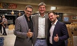 Drei Männer in Anzügen stehen drinnen, geben Daumen hoch. Einer trägt eine Brille, ein anderer hat einen Gürtel. Stühle und Menschen im Hintergrund.