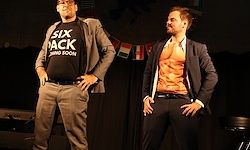 Zwei Männer stehen auf einer Bühne. Einer trägt einen grauen Anzug und ein schwarzes Shirt mit der Aufschrift 'SIX PACK'. Der andere Mann ist ohne Hemd mit einem schwarzen Anzug. Dahinter befindet sich ein schwarzer Hintergrund mit Flaggen.