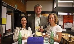 Drei Personen, zwei Frauen und ein Mann, stehen in einem Raum an einem Tisch, lächeln und posieren für ein Foto. Eine Frau hält ein Weinglas, und es gibt Flaschen und eine Box auf dem Tisch.