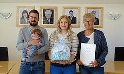 Drei Personen stehen in einem Büro. Ein Mann hält ein Baby, eine Frau einen Korb und eine andere Frau ein Zertifikat. Sie stehen vor einem Schreibtisch und gerahmten Bildern.