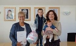 Drei Frauen und ein Kind stehen in einem Raum und halten einen Korb und ein Zertifikat. Hinter ihnen hängen drei gerahmte Fotos an der Wand.