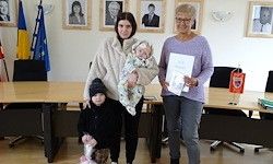 Drei Personen, darunter eine Frau mit einem Baby und eine weitere mit einem Papier, stehen in einem Raum mit gerahmten Bildern und Flaggen an der Wand.
