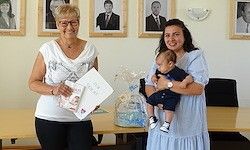 Zwei Frauen stehen vor einem Tisch mit einem Kuchen. Eine hält ein Baby und ein Dokument, während die andere ein Bild und ein Dokument hält. Gerahmte Porträts hängen an der Wand hinter ihnen.