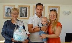 Vier Personen stehen in einem Raum mit einem Baby. Ein Mann hält das Baby, während eine Frau eine Plastiktüte hält. Eine andere Frau trägt eine Brille und hält eine Babydecke. Ein Bilderrahmen hängt an der Wand.