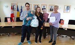 Eine vierköpfige Familie steht in einem Konferenzraum mit zwei Babys, eines in Blau und das andere in Rosa gekleidet. Der Mann und die Frau halten ihre Kinder, während eine Frau ein Zertifikat in der Hand hält. Flaggen und Bilderrahmen schmücken die Wand.