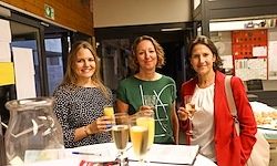 Drei Frauen stehen an einem Tisch mit Gläsern Orangensaft. Die Frau auf der linken Seite trägt ein Blumenkleid. Die mittlere Frau trägt ein grünes Shirt mit weißen Buchstaben. Die Frau auf der rechten Seite trägt eine rote Jacke und hält ein Weinglas.