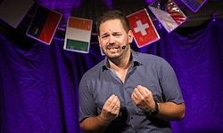 Ein Mann spricht auf der Bühne mit einem Mikrofon. Flaggen verschiedener Länder hängen hinter ihm an einem lila Hintergrund.