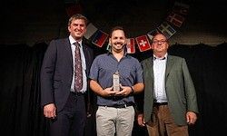 Drei Männer stehen auf der Bühne, einer hält einen Preis. Hinter ihnen zeigt eine Kulisse verschiedene Flaggen, darunter die der Schweiz.
