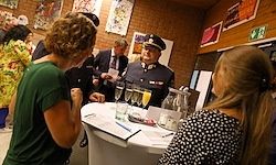 Ein uniformierter Beamter steht hinter einem Tisch mit einem Glas Orangensaft, umgeben von anderen Leuten bei einer Veranstaltung, darunter zwei Frauen, die sich unterhalten.