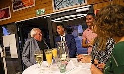 Eine Gruppe von Menschen versammelt sich um einen Tisch mit Getränken, darunter Champagner und Orangensaft. Ein älterer Mann lächelt und schaut in die Kamera. Hinter ihm steht ein Mann im Anzug in der Nähe einer Tür mit Glasscheibe.