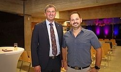 Zwei Männer stehen nebeneinander in einem Innenbereich mit Stühlen und einem Tisch. Der Mann auf der linken Seite trägt einen Anzug und eine Krawatte, der Mann auf der rechten Seite trägt ein blaues Hemd und eine Uhr.