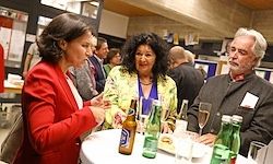Drei Erwachsene, zwei Frauen und ein Mann, sitzen um einen Tisch mit verschiedenen Getränken. Die Frau links trägt einen roten Blazer, die Frau in der Mitte ein gelbes Oberteil und der Mann einen dunklen Anzug.