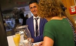 Ein Mann in einem blauen Anzug lächelt, während er an einer Bar sitzt und eine Frau in einem grünen Shirt ansieht. Sie stehen beide vor sich und sehen sich an, mit einem Glaskrug und Gläsern mit Orangensaft auf der Theke.