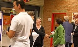 Eine Gruppe von Menschen steht in einem Raum. Eine Frau in einer grünen Jacke schüttelt einem Mann in einem weißen Shirt die Hand.