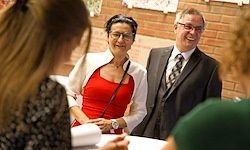 Eine Gruppe von Menschen unterhält sich in einem Raum; eine Frau in Rot und ein Mann im Anzug stehen vor einem Schreibtisch und unterhalten sich mit anderen, eine Ziegelwand im Hintergrund.