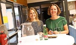 Zwei Frauen sitzen lächelnd am Tisch, mit einer Vase, einer Flasche und einem Stift. Im Hintergrund ist eine Glastür und ein roter Feuerlöscher zu sehen.