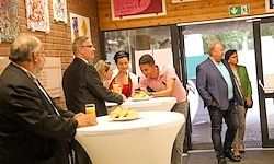 Eine Gruppe von Menschen steht und sitzt um weiße Tische in einem Raum. Ein Mann hält ein Getränk und einen Teller. Andere gehen durch eine Glastür.