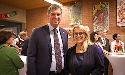 Ein Mann und eine Frau stehen lächelnd bei einer Veranstaltung. Dahinter unterhalten sich Leute an Tischen und in der Nähe einer mit Gemälden geschmückten Ziegelwand.