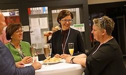 Eine Gruppe von Menschen sitzt um einen Tisch mit Getränken und Essen. Eine Frau mit Brille spricht lächelnd zu anderen. Sie scheinen eine gesellige Zusammenkunft zu genießen.