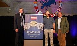 Drei Männer stehen vor einem Banner mit dem Rotary Club Logo und Flaggen hinter ihnen. Ein Mann trägt einen schwarzen Anzug, einer ein graues Shirt und der andere einen grünen Mantel.