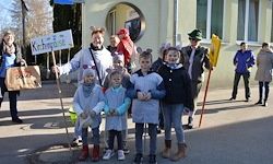 Eine Gruppe von Kindern in Kostümen posiert für ein Foto vor einem Gebäude, ein Mann hält ein Schild und einen Besen, und Menschen stehen im Hintergrund.