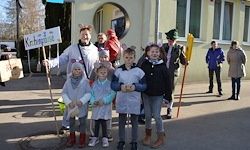Eine Gruppe von Kindern in Kostümen, darunter ein Mann in einem Bärenkostüm, posiert für ein Foto auf der Straße, mit einem Gebäude und Menschen im Hintergrund.