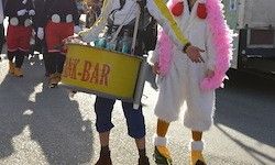 Zwei Menschen in Hühnerkostümen, einer trägt einen gelben Korb mit der Aufschrift 'Snack-Bar', laufen in einer Parade.