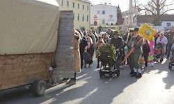 Eine Gruppe von Menschen geht auf einer Straße, einige in Kostümen, trägt Pflanzen und Flaggen, mit alten Wagons und Gebäuden im Hintergrund.