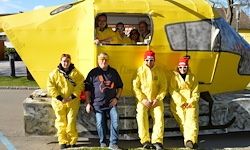 Fünf Personen in gelben Schutzanzügen posieren vor einer gelbfarbenen helikopterartigen Struktur, wobei zwei andere im Inneren durch die Fenster schauen.