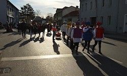 Eine Gruppe von Menschen in Kostümen geht eine Straße entlang, mit Gebäuden und Bäumen auf beiden Seiten. Die Schatten der Menschen fallen auf die Straße.