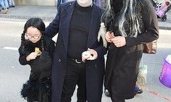 Eine Familie in Halloween-Kostümen steht auf der Straße, ein Kind hält ein Stück Süßigkeit, während Erwachsene Schwarz tragen mit langen Haaren und einem Mantel.