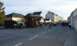 Ein Traktor ist auf einer Straße geparkt, mit einem mit Heu dekorierten Festwagen in der Mitte. Viele Menschen gehen auf der Straße und dem Bürgersteig. Es gibt Häuser und ein Gebäude auf beiden Seiten der Straße.