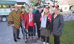 Eine Gruppe von Menschen in verschiedenen Kostümen steht auf einem Bürgersteig vor einem Gebäude. Eine Frau benutzt einen Gehwagen.