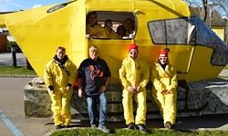 Fünf Personen in gelben Anzügen posieren vor einem großen gelben Hubschraubermodell, während einige andere darin sitzen. Die Szene spielt sich auf einem Rasenstück ab.